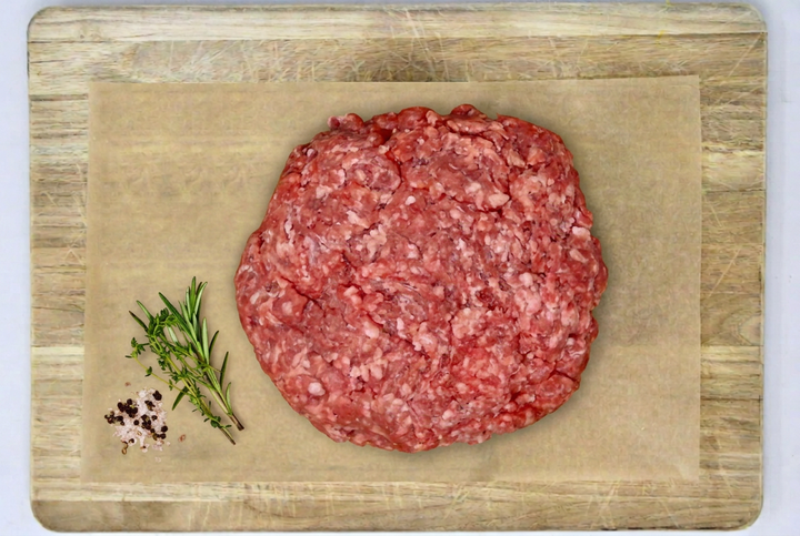 Raw grass-fed ground beef organ blend patty placed on parchment paper atop a wooden cutting board, with a sprig of rosemary and coarse salt on the left side.