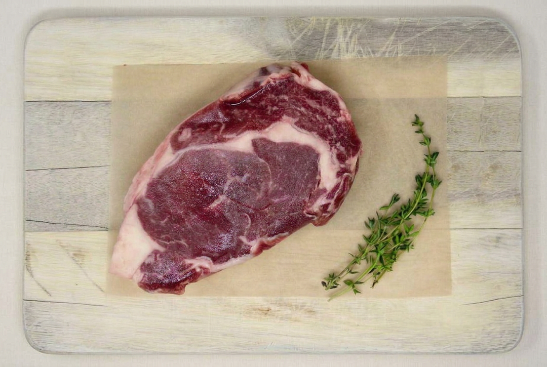 Raw boneless beef ribeye steak with thyme on a wooden cutting board with its outline visible.