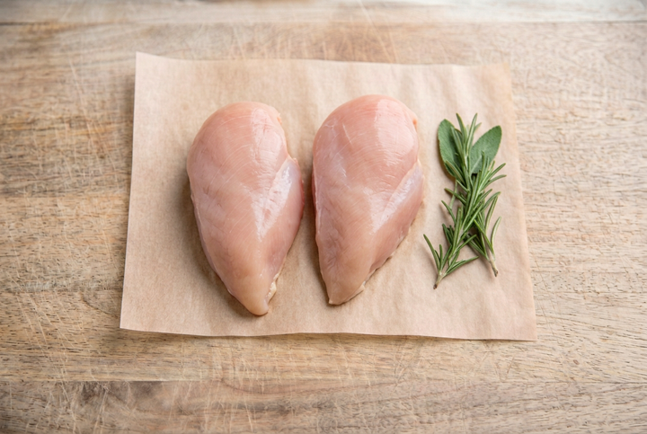 Two raw boneless skinless chicken breasts on parchment on a wooden cutting board beside fresh herbs.