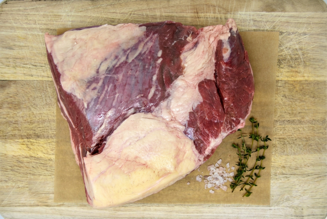 A raw cut of grass-fed beef brisket with fat cap, placed on a sheet of brown butcher paper atop a wooden cutting board, with coarse salt and a sprig of fresh thyme to the right.