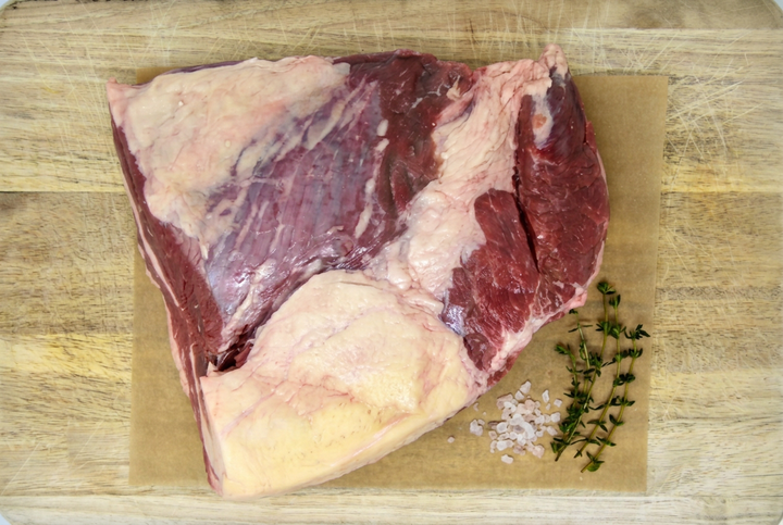 A raw cut of grass-fed beef brisket with fat cap, placed on a sheet of brown butcher paper atop a wooden cutting board, with coarse salt and a sprig of fresh thyme to the right.