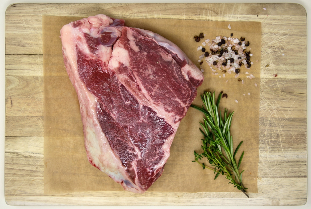 Raw beef chuck roast with herbs and seasoning on a wooden cutting board with its outline visible.