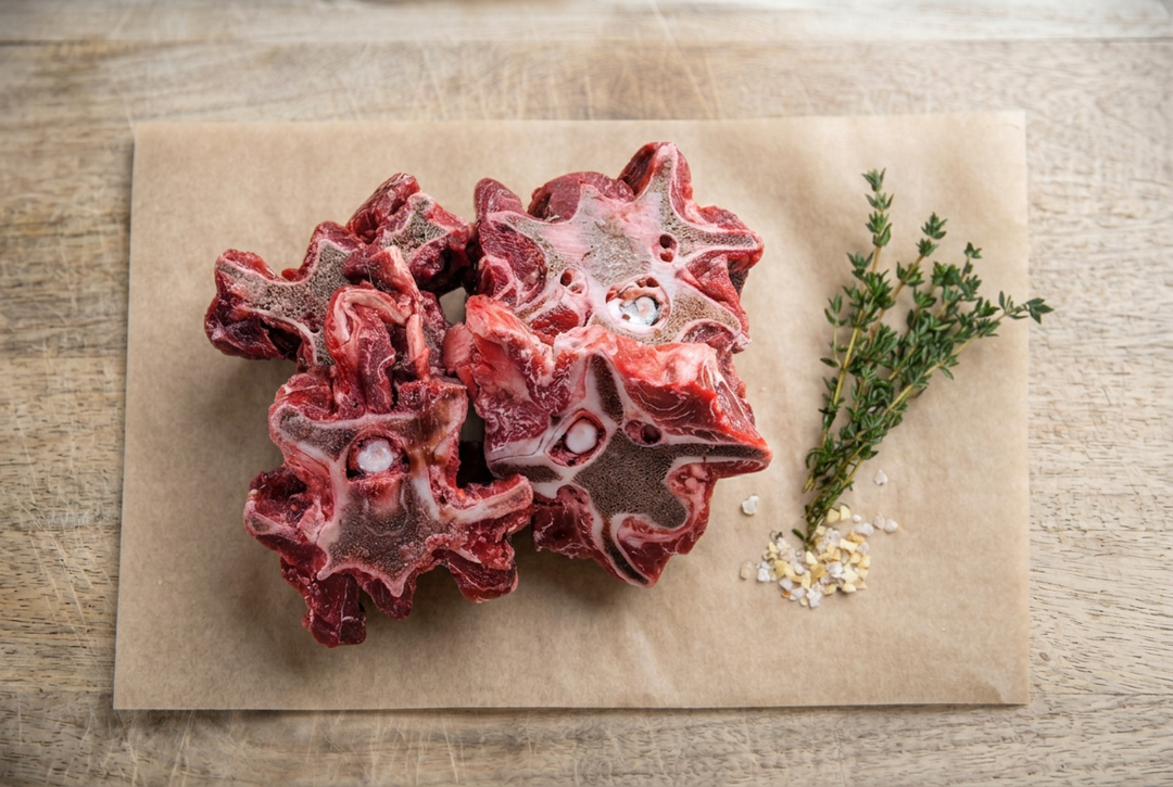 Raw beef meaty neck bones on parchment on a wooden cutting board with herbs on the right.