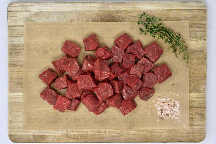 Chopped raw grass-fed beef tenderloin filet tips arranged on parchment paper atop a wooden cutting board, with a sprig of thyme and coarse salt nearby.