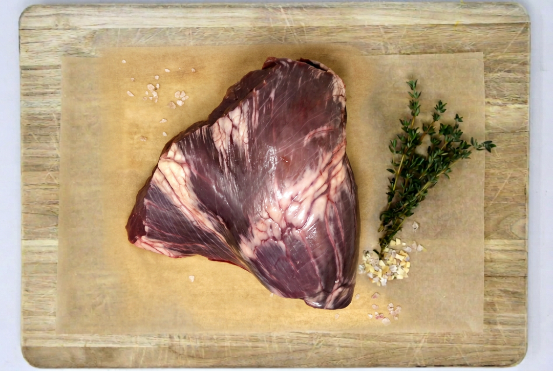 A raw cut of grass-fed beef heart with visible marbling, placed on parchment paper atop a wooden cutting board, with a sprig of herbs and coarse salt nearby.