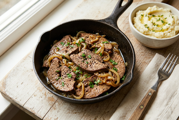 A cast iron skillet filled with sliced grass-fed beef liver and caramelized onions, garnished with chopped chives, set on a rustic wooden board next to a bowl of mashed potatoes and a fork.